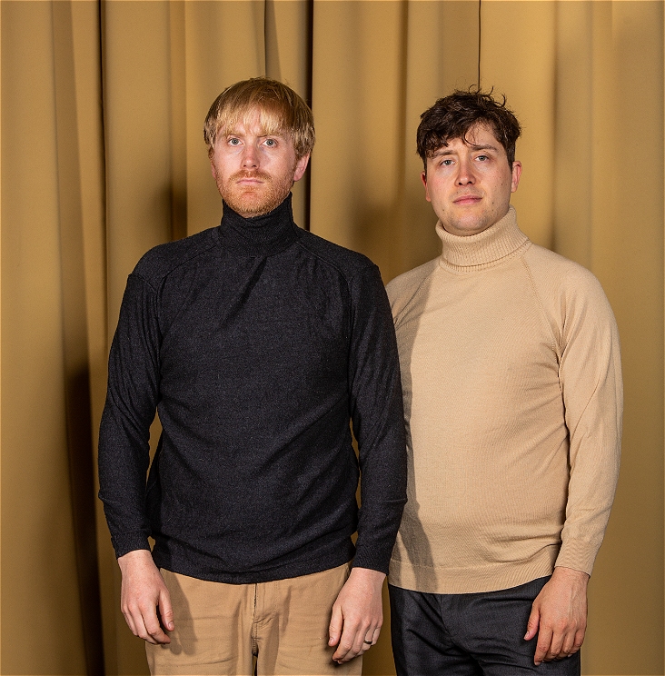 Two people stand together - they have short hair - one has a dark top and light brown pants, the other has dark pants and a light brown top.