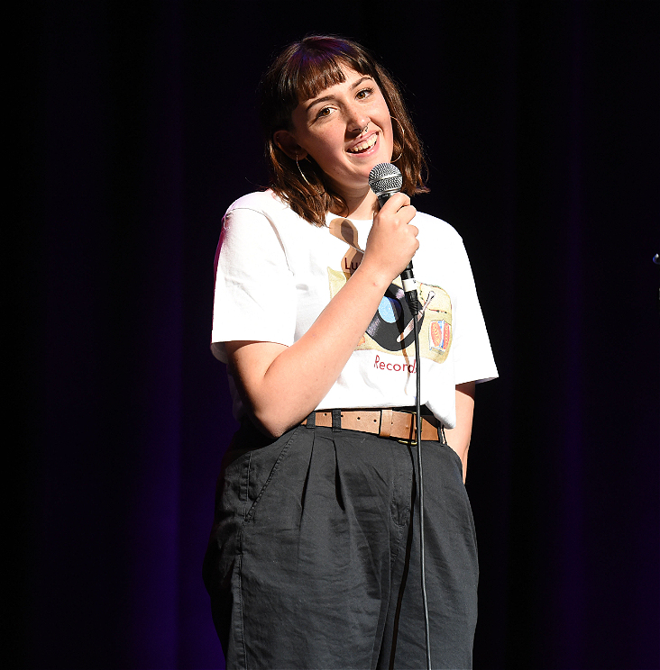 A girl with brown hair and a smile on her face stands holding a microphone with one hand. She wears a white tshirt and hight-waisted black pants with a brown belt.