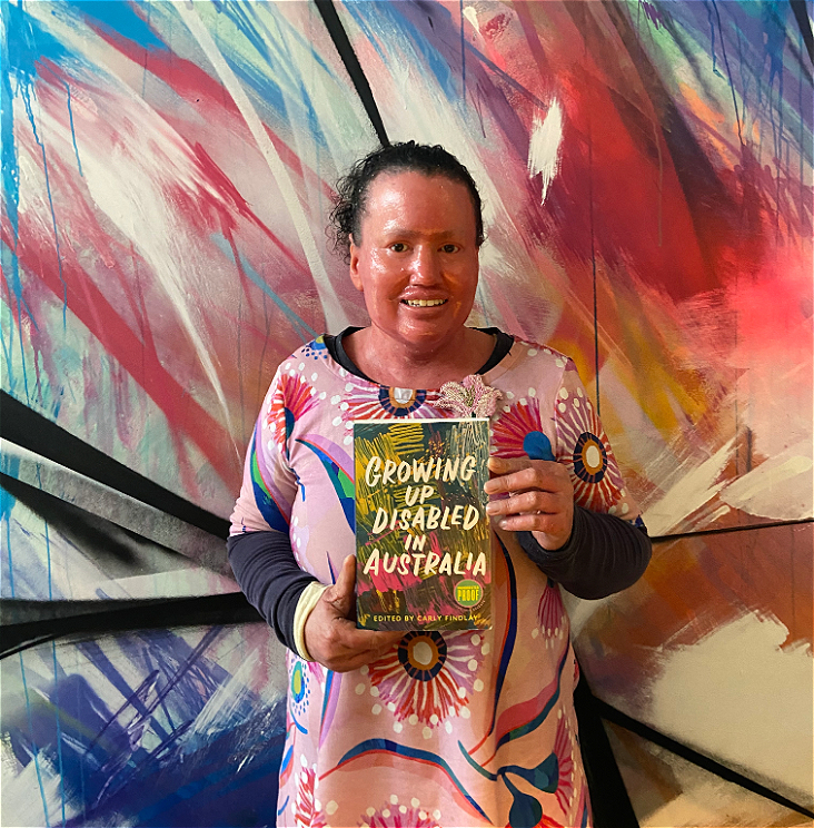 A women holds a colourful book, wearing a colourful dress in front of a colourful background. The colour palette is blues, pinks and purples, there is an floral theme. The book title reads "Growing Up Disabled in Australia"