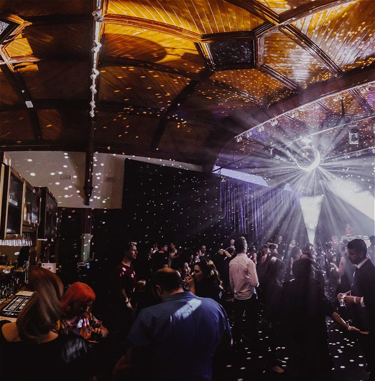 A room lit by a very shiny disco ball is full of people chatting and dancing. There is a bar in the left of the image serving drinks. The room looks old and beautiful with black curtains on the walls.