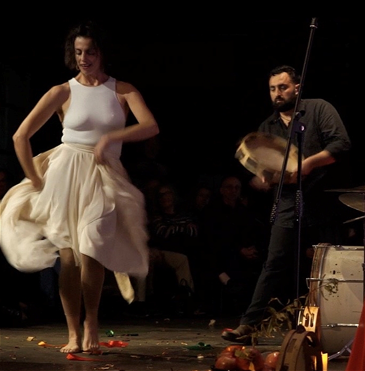 A woman in a white dress dances and her skirt twirls around her. Behind her a man wearing black hits a tambourine.