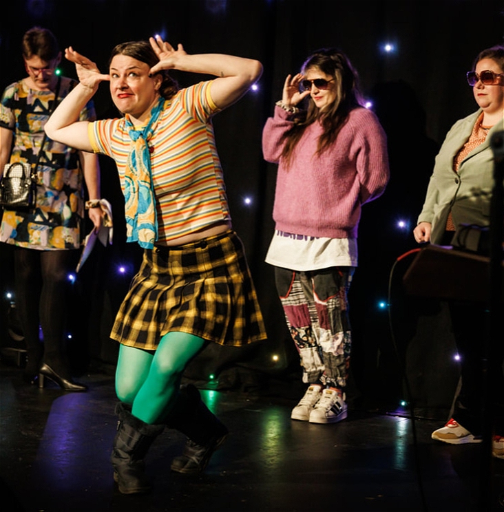 A person wearing colourful clothing stands on a stage. The floor of the stage is black, and there is a black curtain behind them, upon which are sparkling fairy-lights which resemble stars.They are wearing a striped shirt, a plaid yellow skirt, and vibrant green tights. They are pulling a credulous face and hold their hands up to their temples in a mocking gesture. Behind them three people stand. They wear dark sunglasses and look directly ahead.