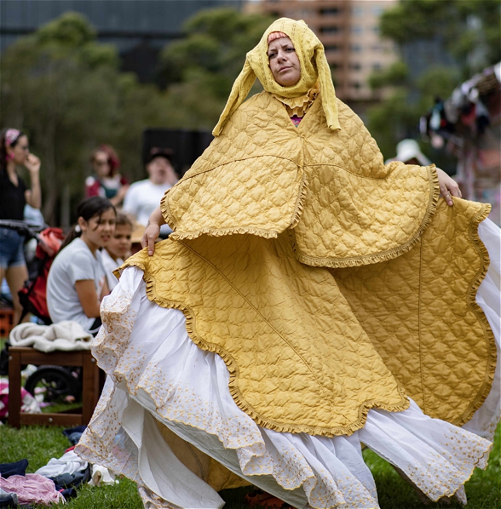 a person wearing a bed spread as a dress walks on grass. there is some audience in the background watching
