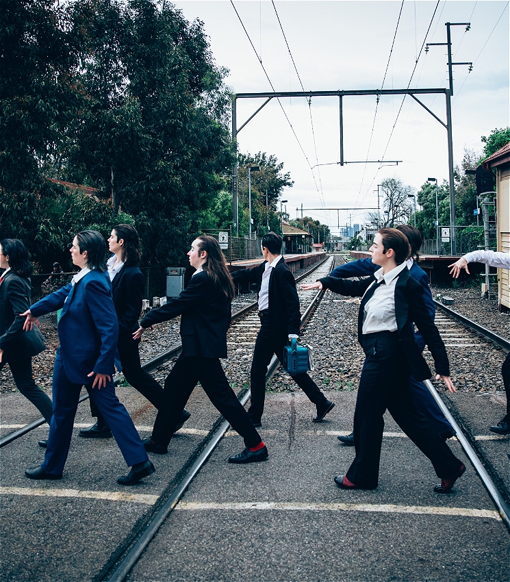 Eight people in wearing suits cross the train tracks. Their hair is slicked back and they walk in unison.