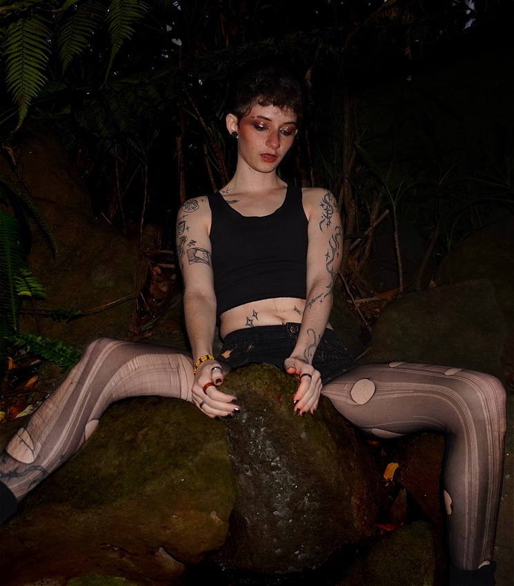 A trans masc person with smokey makeup and tattoos down their arms. They wear a black binder, shorts and ripped tights straddles a large mossy rock. They are surrounded by rainforest.