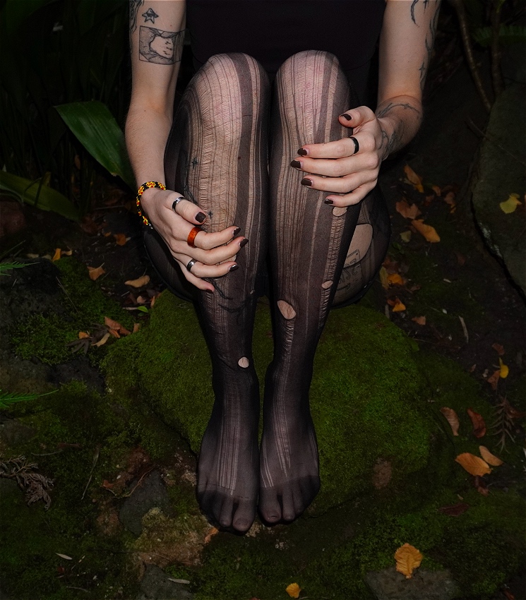 A person sits and holds their knees to their chest on the mossy forest floor. They are wearing ripped black tights on their legs, lots of rings on their hands, and have tattoos across their arms.
