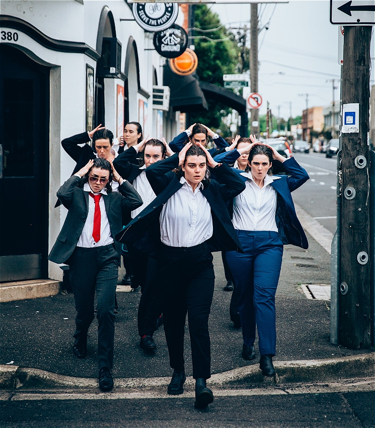 A large group of people dressed in suits stride towards the camera. They are all slicking back their hair.