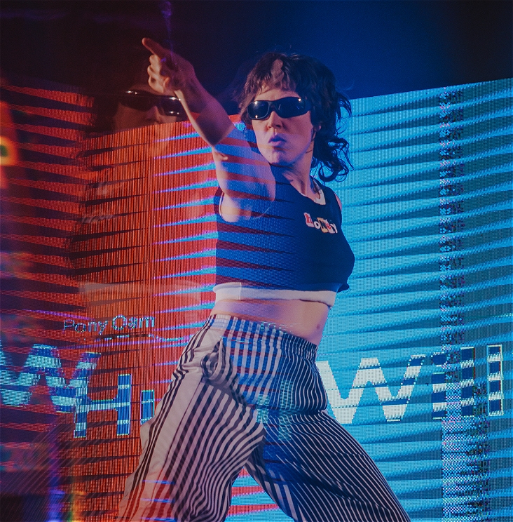 A performer with a brown mullet is pointing their finger. They're wearing black sunglasses, a black crop top and black and white stripped pants