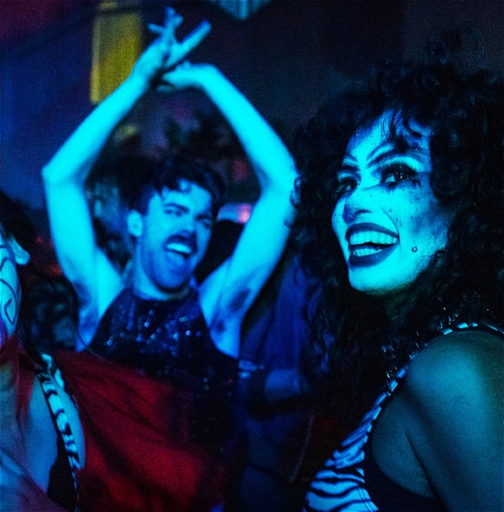 Two people on a dancefloor. One of them has bold, clown makeup on. The second has their arms raised in joy