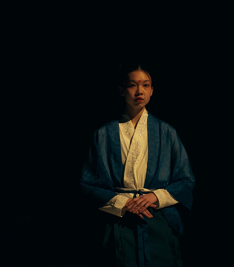A Chinese dancer stands to the right of the frame, wearing traditional Han-style clothing. She is lit with warm side lighting that highlights her face and garments against a black background. Her expression is stern and composed. Her arms are held regally in front of her waist. Her makeup includes a traditional Chinese huadian - a decorative flower mark placed on her forehead.