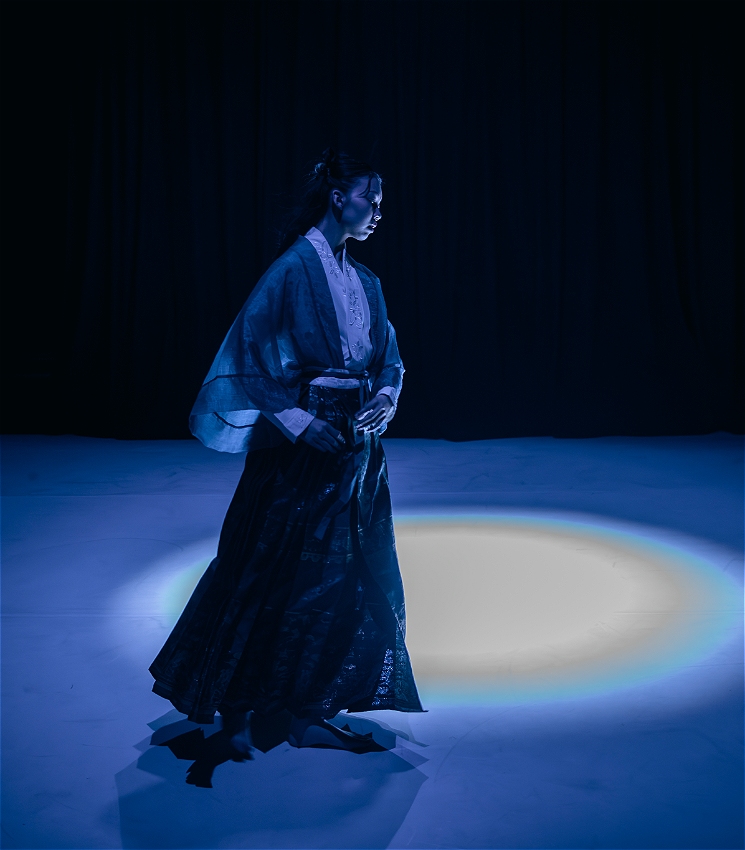 A full-body image of a Chinese dancer captured in profile, mid-step. She wears traditional Han-style clothing, including a flowing skirt and a long-sleeved top with wide sleeves. Her body tilts slightly as she walks toward the edge of the frame, angled toward a soft blue spotlight at the centre. The light catches the edge of her garments, creating contrast against the dark background.