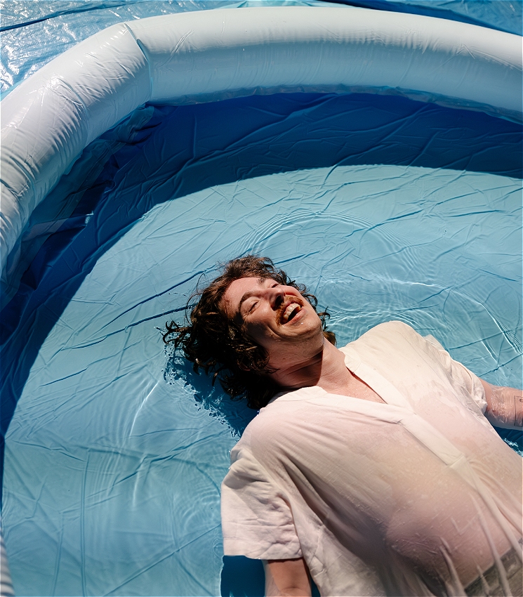 A photo of a young man lying on his back. He is half submerged in a blue blow up pool, his white clothes are wet and semi-transparent. His hair is brown and curly and spreads out in rays from his head, floating on the surface of the shallow water. He has an enormous smile, like he's been caught in the photo mid belly-laugh.