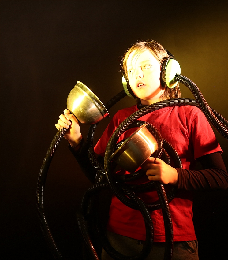 A child in a darkened space with a curious expression holds a unique instrument connected via thick tangled tubes to the strange headphones they are wearing and listening to.