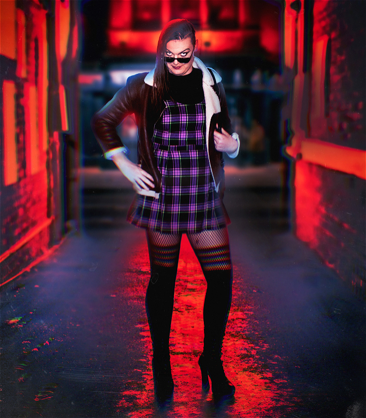 The image features Courtney Maldo (a perth based transwoman comedian) standing alone in an alley at nighttime. She's wearing a purple and black tartan schoolgirl dress and a large brown winter jacket. She's wearing 5 inch heels and tights. She has sunglasses perched on her nose and is staring into the soul of the viewer.