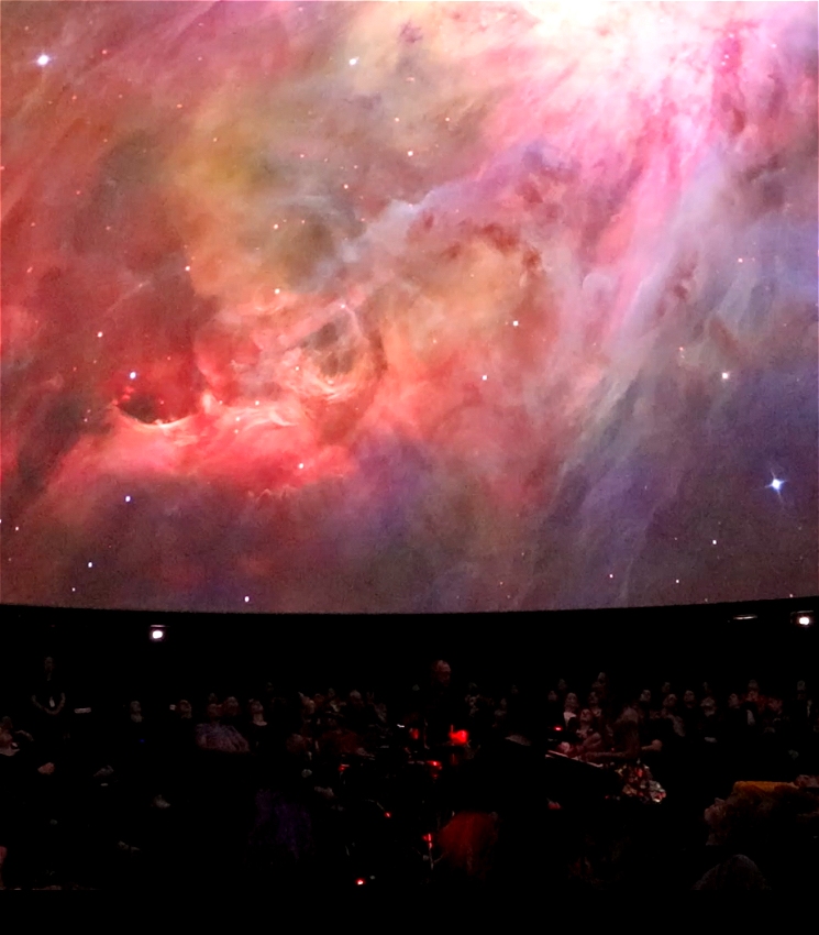 A string quartet perform in a dark setting, their faces and instruments illuminated by the amber glow of the iPads they are reading music from. Above them, dramatic pink clouds swirl across the domed ceiling of the planetarium.