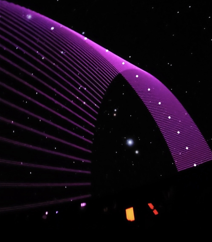 A string quartet perform in a dark setting, their faces and instruments illuminated by the amber glow of the iPads they are reading music from. Above them, a giant purple arch soars across the domed ceiling of the planetarium.