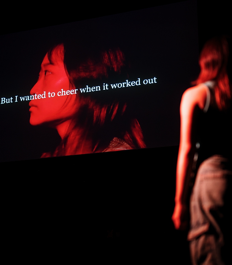 A person faces away from the camera and looks up at a screen where an image of their face is projected. On the screen, there is text that reads: But I wanted to cheer when it worked. The rest of the room is in darkness.