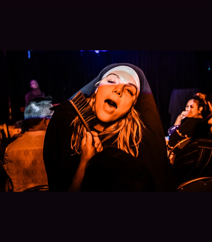 A woman dressed as a nun faces the camera with a look of ecstasy on her face while someone brushes her jaw with a hand-held broom. In the background, audience members can be seen looking at her in shock and amusement.