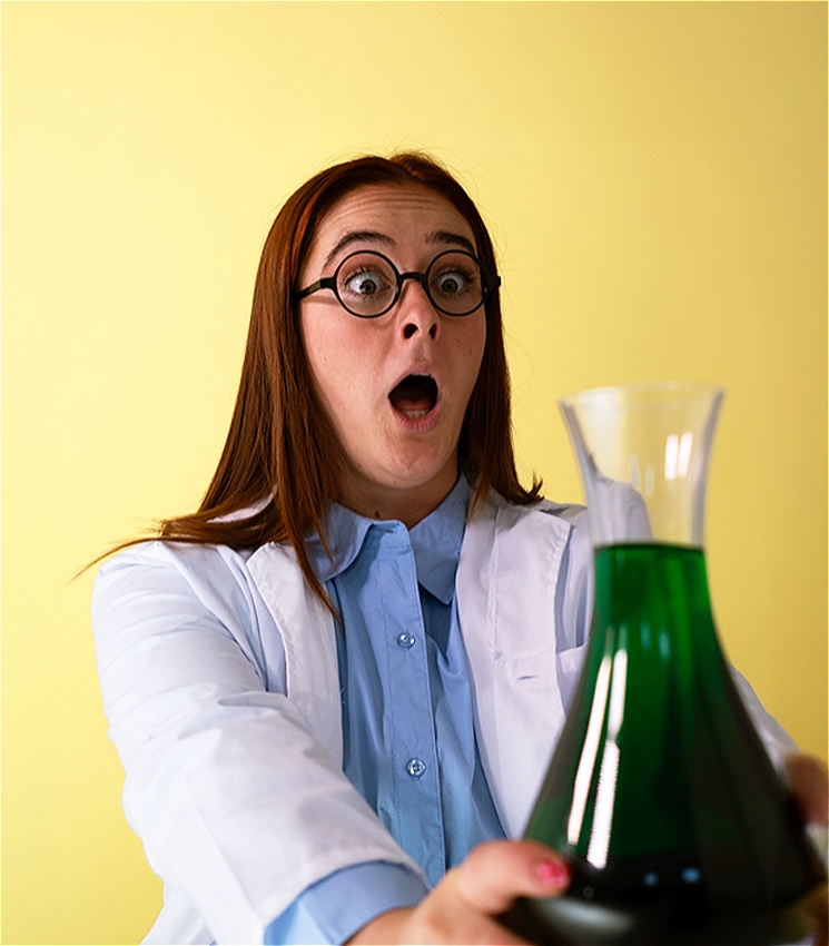 A young woman in a lab coat is holding a large flask filled with bright green liquid. She looks completely stunned – like something just happened that she didn’t expect. Her eyes are wide, her mouth is open in shock. She’s wearing round glasses and has long, red hair. The background is a bright yellow.