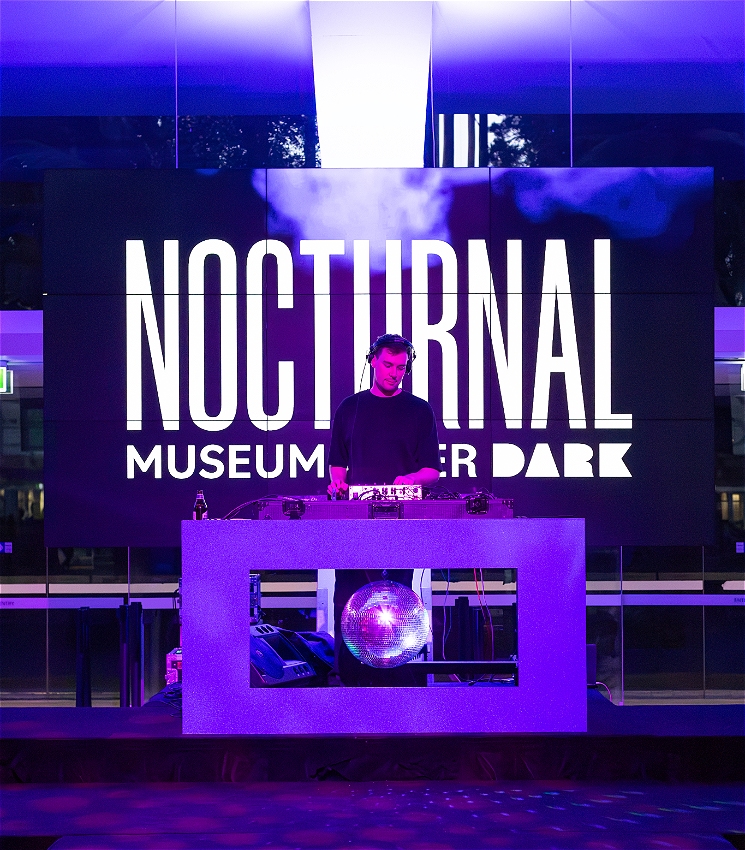 DJ Boyd Kelly plays music on DJ decks in front of a digital sign that reads: Nocturnal Museum After Dark. There is a disco ball beneath the DJ decks.