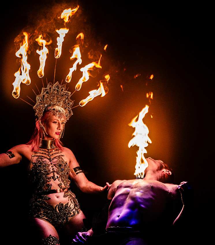 A woman wearing a flaming headdress and a bejeweled bodice looks down on a shirtless man who sits, leaning back in a chair, with his chest on fire.