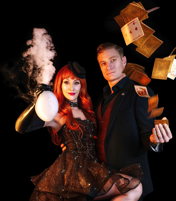 A handsome man throws cards while smouldering at the camera, and a very well dressed young woman smile, holding a bubble that is pouring smoke like a volcano