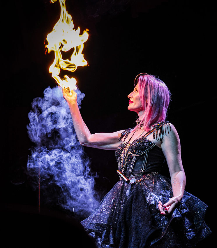 A woman in a glimmering black skirt looks up and to the left as a gout of flame that seems to erupt from her hand, as smoke billows around her