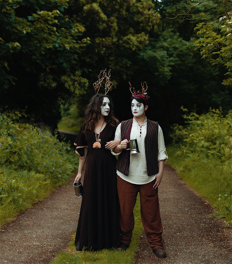 Two women stand on a grassy path while arm in arm and looking at each other. They both hold metal tankards and wear antlers on their heads. There is a pathway leading to a tunnel of trees in the background.