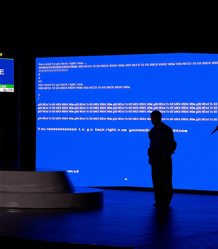 A person stands on stage in front of a large blue screen filled with glitchy text repeating “You need to go back right now.” The lighting is dark, and the mood feels tense and mysterious. The set is minimal, with a white platform and a few plants.