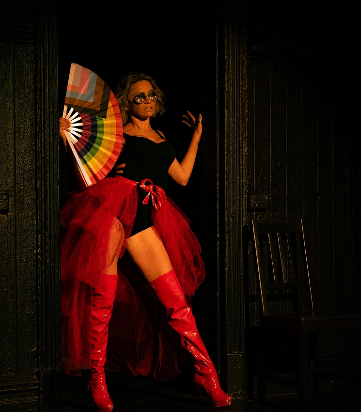 Dr. Jae West poses on stage with a pride flag handheld fan, big red boots and a red skirt.