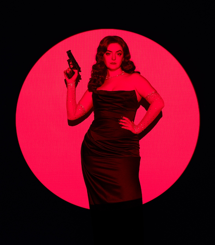 A red spotlight illuminates Abbey in a black room. She's standing with one hand on her hip and one holding a rhinestoned prop gun. She is wearing a black strapless dress, red lipstick, vintage brunette waves and sparkly gloves.