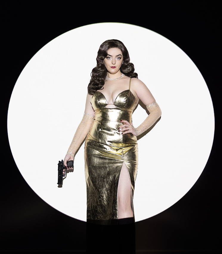A bright spotlight illuminates Abbey in a black room. She's standing with one hand on her hip and one holding a rhinestoned prop gun. She is wearing a metallic gold dress with a thigh high split, red lipstick, brunette waves and sparkly gloves.