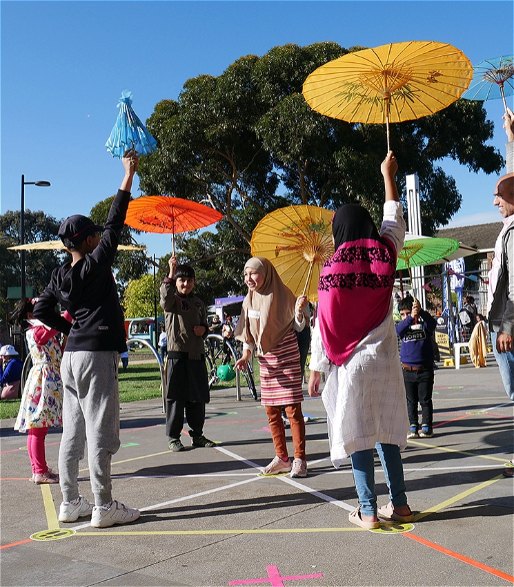 A lively outdoor scene where children and adults, many from diverse cultural backgrounds, joyfully participate in a playful public art activity. They hold vibrant, colorful paper umbrellas while standing on a ground marked with bright geometric tape.