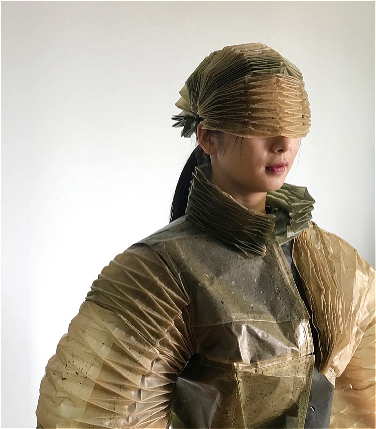 Artist Yajuan Cheng stands against a plain white background wearing an intricately pleated, translucent outfit in earthy tones of brown and green. Her eyes are covered by a visor-like pleated headpiece, and her jacket features exaggerated folds and texture.
