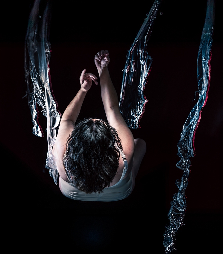 A dark-haired person in a nude bodysuit is falling backwards with their back and the top of their head in the middle of the image. Their face is not able to be seen. There are tendrils of blue light that almost look like water cascading vertically down the image.