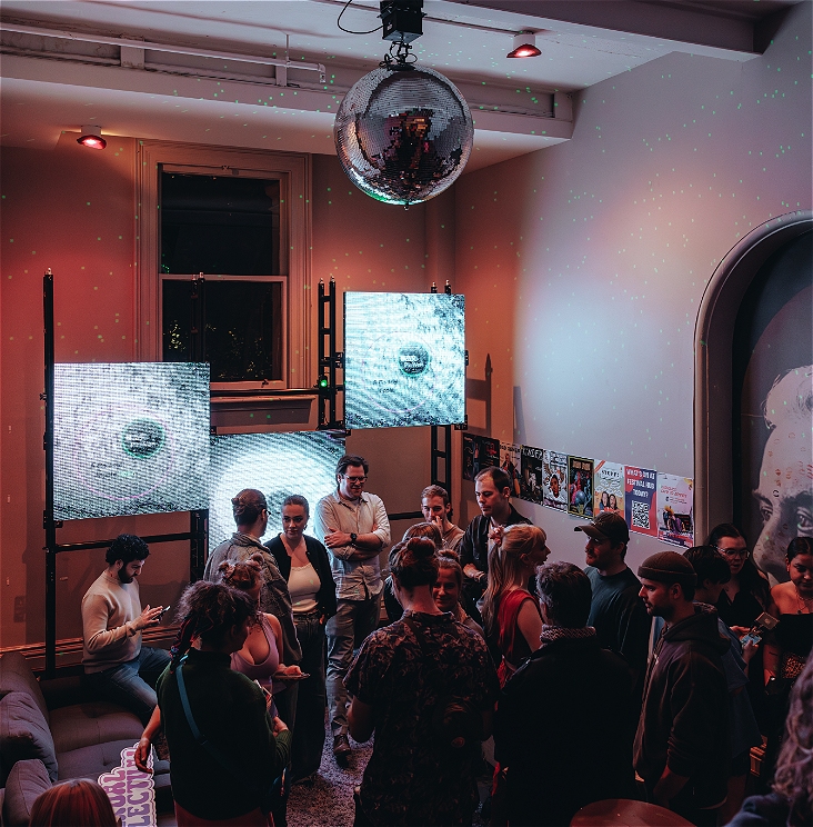 A crowd stands mingling in the hallways of Trades Hall, there are white pixapixelatedted LED screens in the background. Everyone looks happy and social.