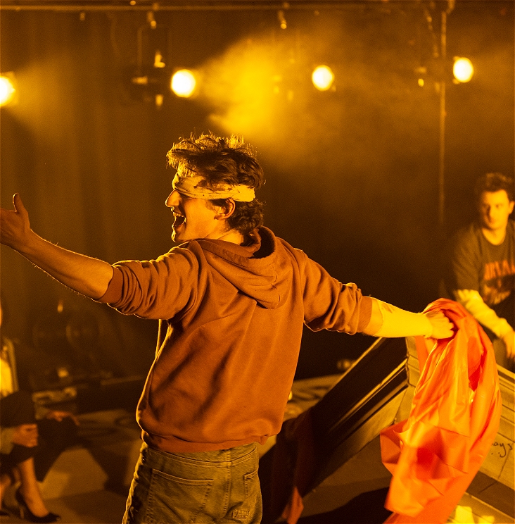 Someone blindfolded stands with their back to the camera and their arms outstretched in exclamation. There are theatre lights and set in the background casting a warm light over everything.