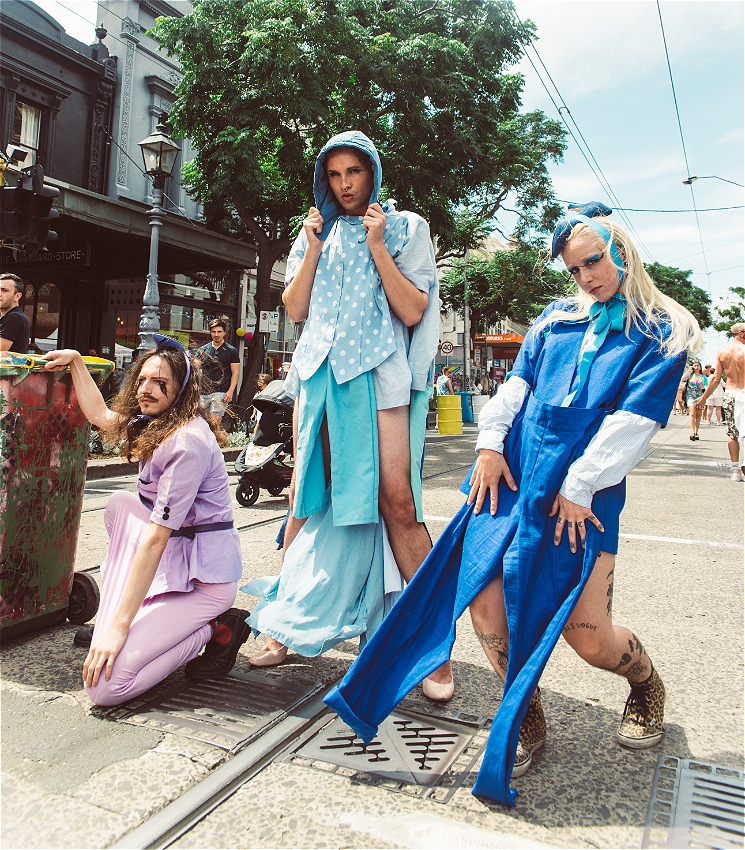 Three adults pose dramatically in the middle of a city street, wearing colourful, DIY-style outfits made from upcycled fabric. One wears lavender and kneels beside a bin, another stands in blue layers with a hood, and the third strikes a bold pose in bright cobalt blue. The background shows pedestrians, tram tracks, and city buildings under a clear sky.