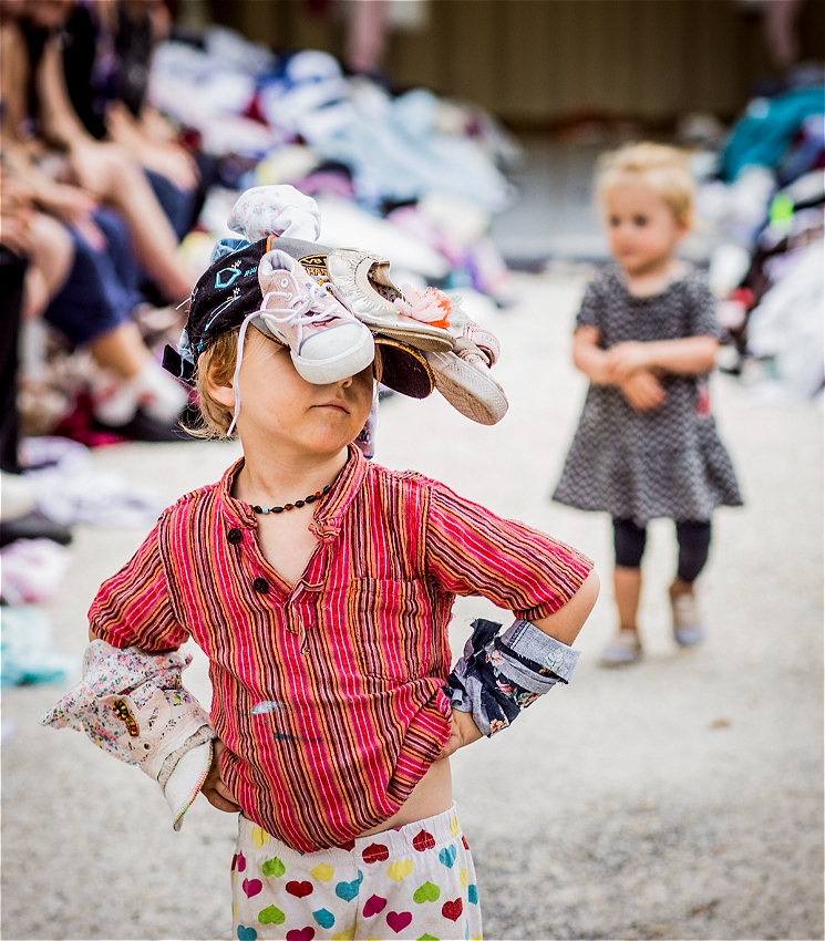 A young child stands confidently on a makeshift runway, hands on hips, wearing a striped red shirt, heart-print pants, and a headpiece made of hats and shoes. Their expression is proud and playful. In the background, another small child watches while adults sit along the runway edges, smiling.