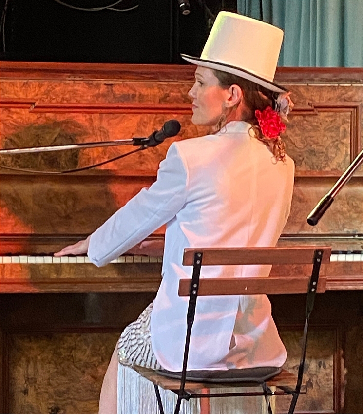 Lead performer Marie-Thérèse wears a white top hat with white tuxedo jacket over a 1920's flapper dress, sitting at an upright piano with a microphone, indicating she has been singing. Her head is turned to the left, presumedly towards the audience, whilst her hands remain on the piano, presumedly playing music.