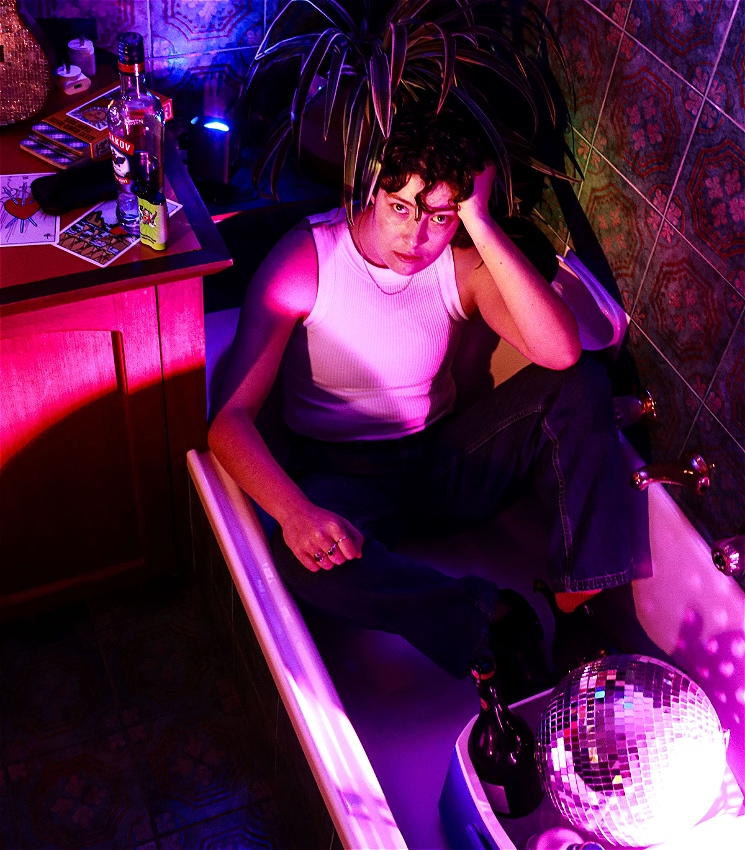 A non-binary person with short curly hair and wearing a tank top sits in a bathtub looking broodingly up at the camera. The scene is lit by fluorescent purples and blues, and in the bathtub is an eski with a disco ball and bottles of alcohol. On the counter next to the tub is strewn vapes, tarot cards, and more alcohol.