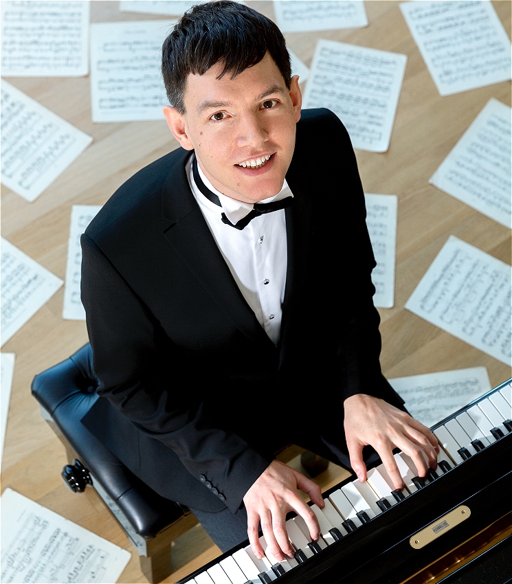 An image taken from a raised place with the camera looking down at Matthew Shiel playing the piano. Matthew - a white man with short black hair and a shaven face - is wearing a tux and black bow-tie, while looking up to the camera and smiling. Both his hands are holding down a chord on the white keys of a piano, while piano scores rest all around the background of the image.