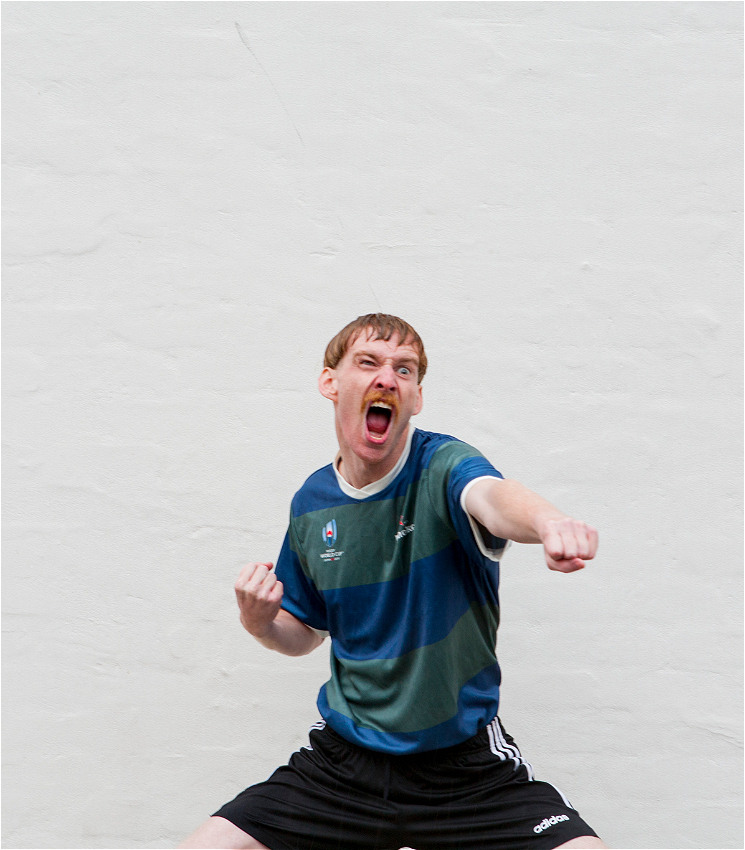 A rambunctious footy hooligan punches the air screaming. He has a bad bowl cut and a shitty moustache but a lot of self confidence. He wears a blue and green striped shirt and black sports shorts. He is standing in front of a white wall.