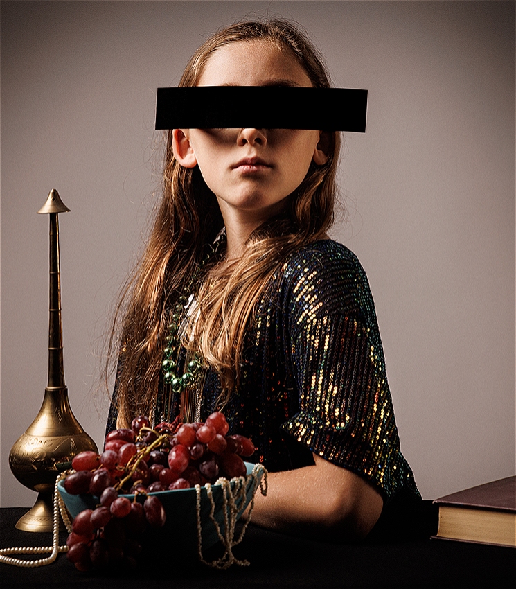 The image shows a child sitting at a table adorned with a gold vase, a bowl of purple grapes, pearl jewellery and a book. They have long golden hair draped over their shoulders and they are wearing a dark green and gold sequin shirt with a green beaded necklace. Their eyes are covered with a black rectangle or censorship bar so that their identity is hidden.