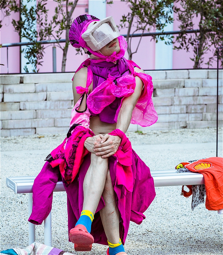 A person sits cross-legged on a bench, dressed in a dramatic magenta costume made from textured fabric, with a large head wrap and a soft, box-like mask covering their eyes. They wear mismatched socks and pink Crocs. Behind them is a pink building and some greenery, creating a surreal, theatrical contrast.