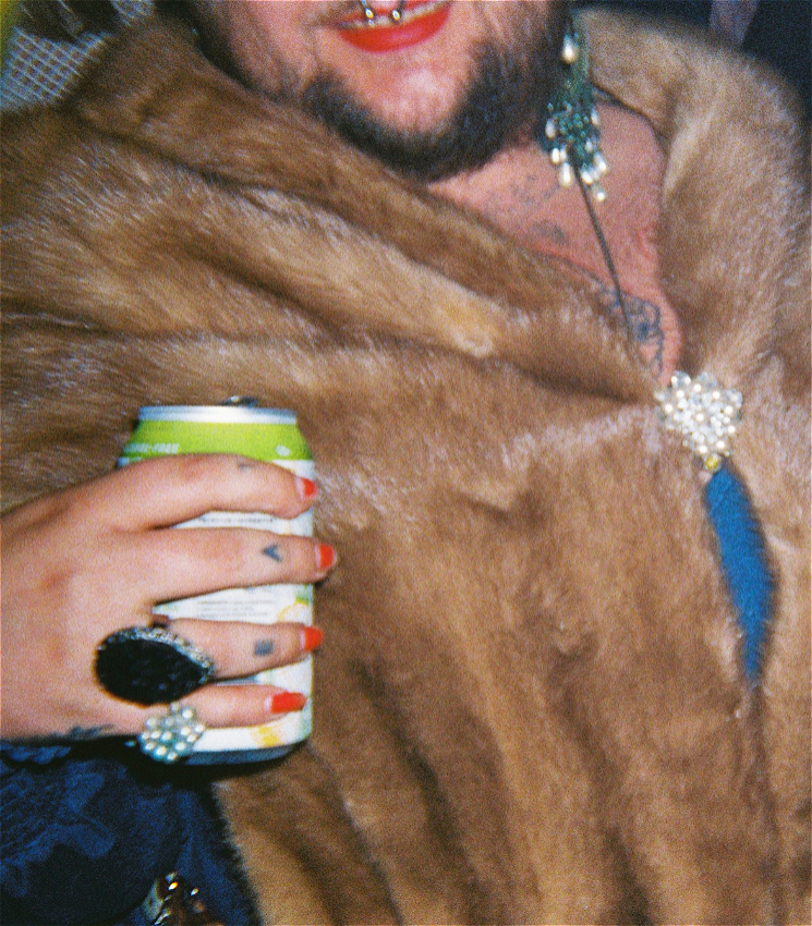 A person with a beard, red lipstick and a septum piercing, is wearing a brown fur coat and holds a can in a hand with big red nails.