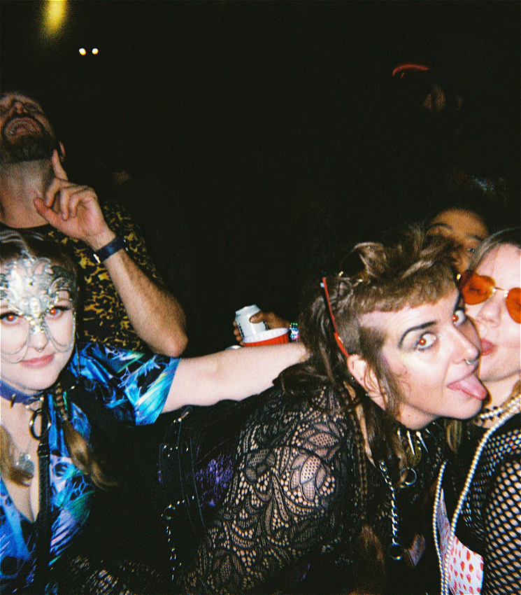 Four peoples are entangled in a playful drunken dance. bodies are entangled in a playful drunken dance. The person in the middle wears black lace, has cropped hair and is sticking their tongue out while looking at the camera. The person next to them wears a silver mask over their eyes.