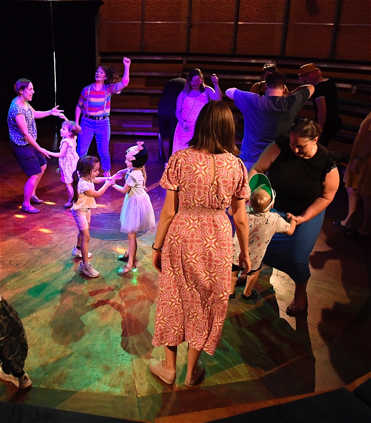 Adults and children dancing together under disco lights. Hana Fiesta in rainbow shirt and blue pants dances with them.
