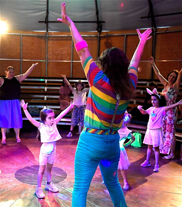Hana Fiesta in rainbow shirt and blue pants leads a dance for children and adults, they all have their hands in the air