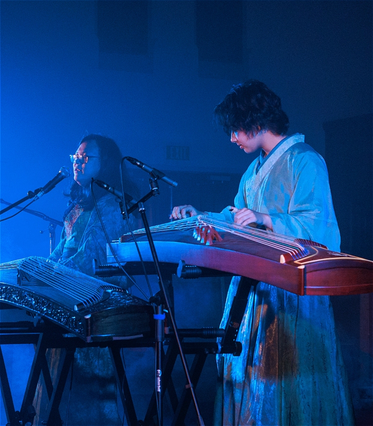 Two musicians perform onstage in a dimly lit venue with high ceilings and wooden panelled walls. They wear flowing, traditional-style garments, and one musician on the right plays guzheng (a traditional Chinese zither), the musician on the left is playing a keyboard (though you can not see the keyboard as it's hidden by the guzheng in front of it). A blue stage light casts a soft glow and haze around them, creating an ethereal atmosphere. Microphones and cables surround their setup, and the audience is partially visible in shadow on the left. The mood is calm, contemplative, and immersive.